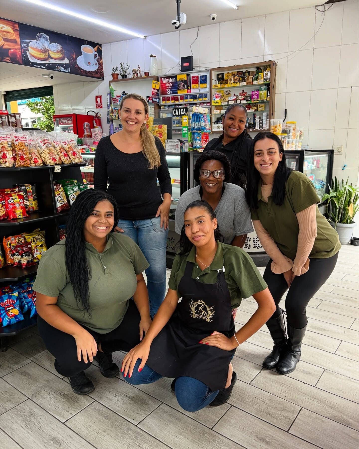 Seis funcionárias da padaria posando juntas dentro da loja, de uniforme e avental com o brasão