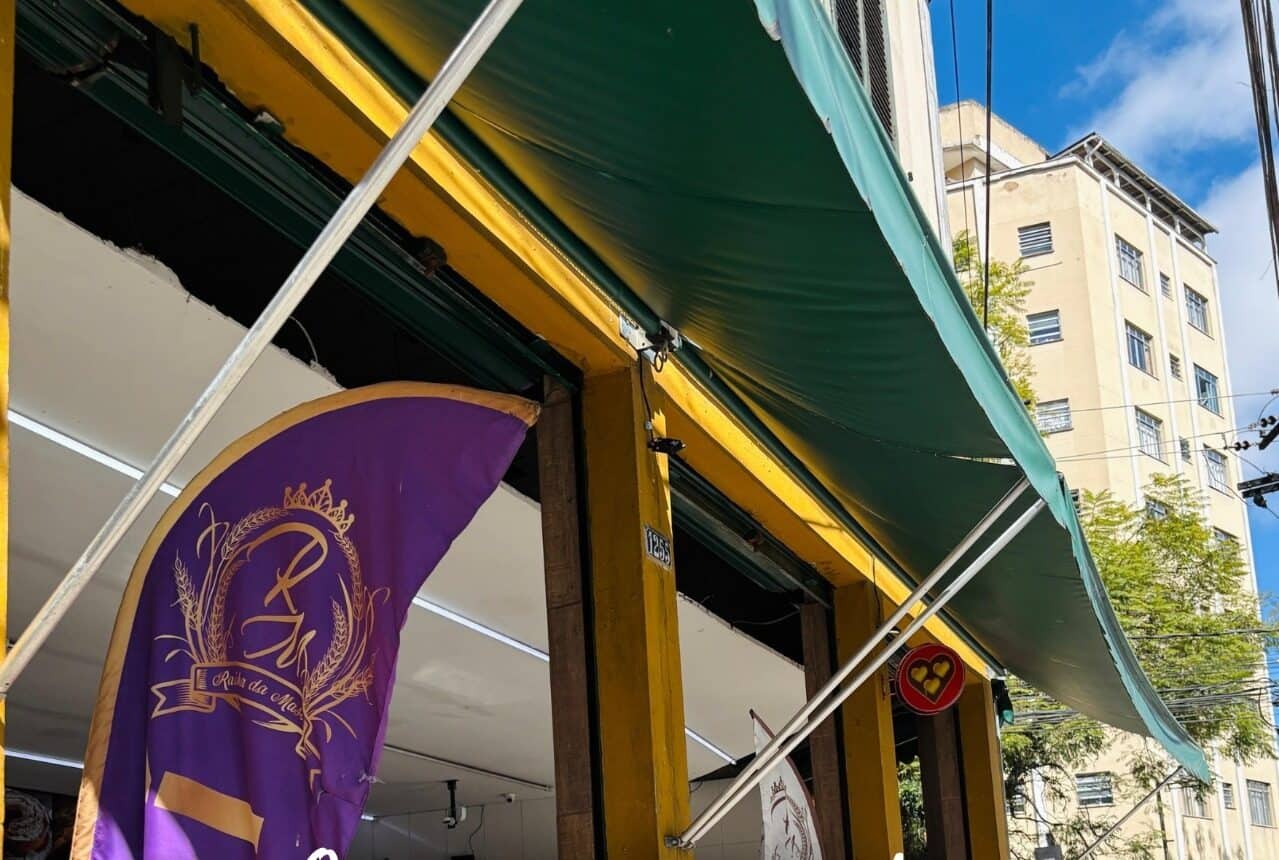 Fachada da padaria, com toldo verde e a bandeira roxa com o brasão da Rainha da Massa