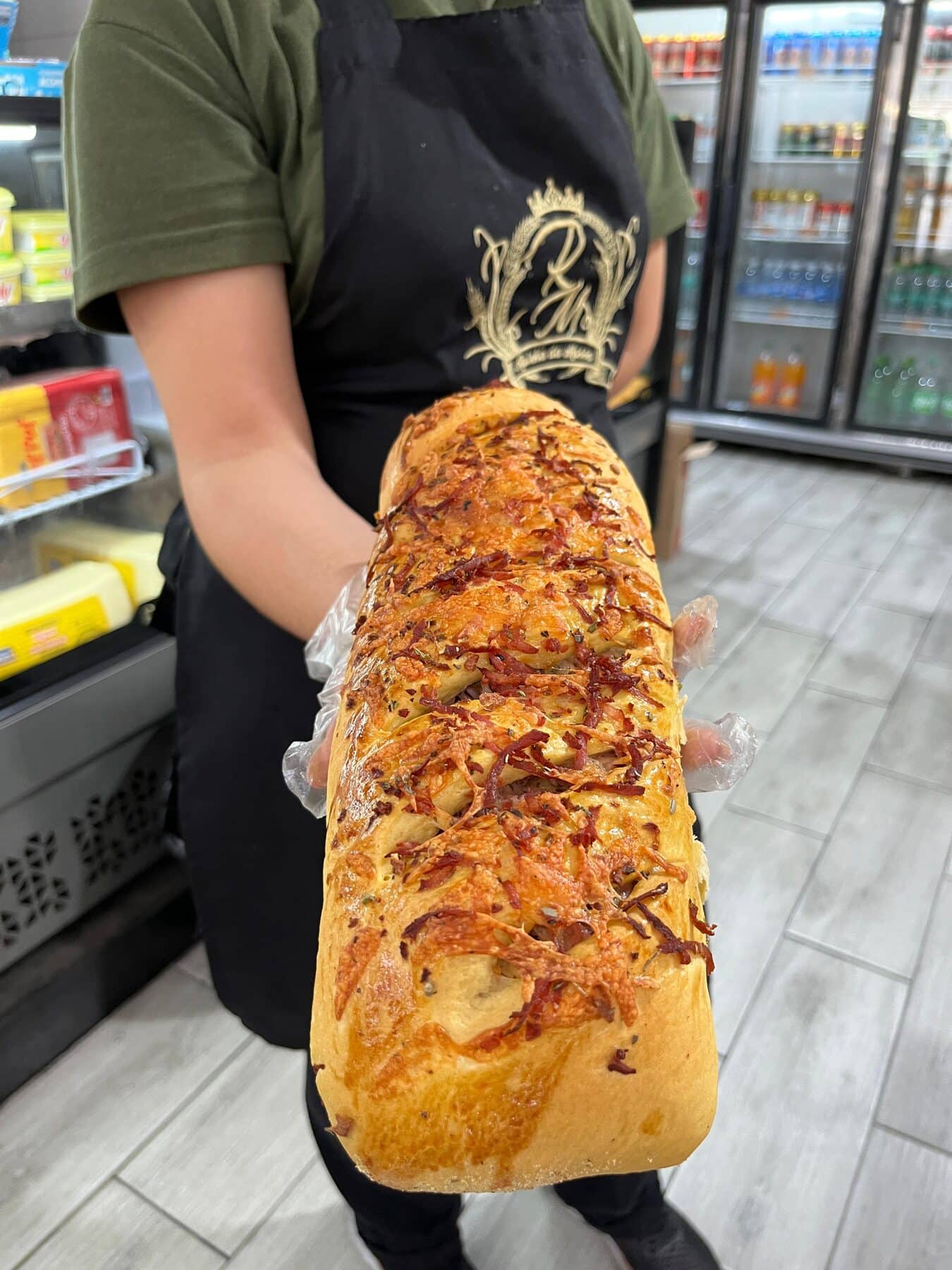 Atendente com o avental do brasão da padaria segurando um pão recheado do tamanho do antebraço