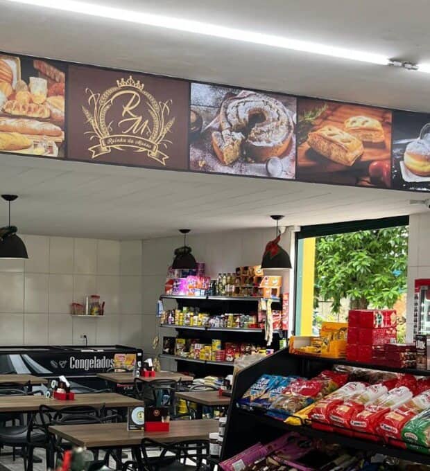 Salão da padaria, com mesas, balcão e o painel de produtos com o brasão na parede