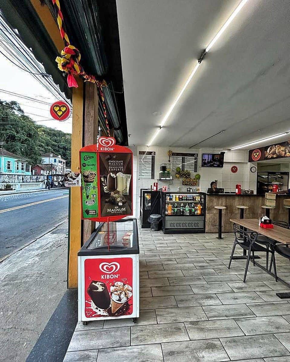 Entrada da padaria vista de dentro, com o freezer de sorvete, as mesas altas e a rua de Petrópolis ao fundo